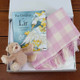 Foxford Baby Blanket Gift Set-Pink. A gift box contains the book "The Children of Lir" by Laura Ruth Maher, a Foxford soft pink and white checkered blanket, and a small plush teddy bear. The items are neatly arranged on a wooden surface. Foxford Baby Blanket Gift Set-Pink. A gift box contains the book "The Children of Lir" by Laura Ruth Maher, a Foxford soft pink and white checkered blanket, and a small plush teddy bear. The items are neatly arranged on a wooden surface.